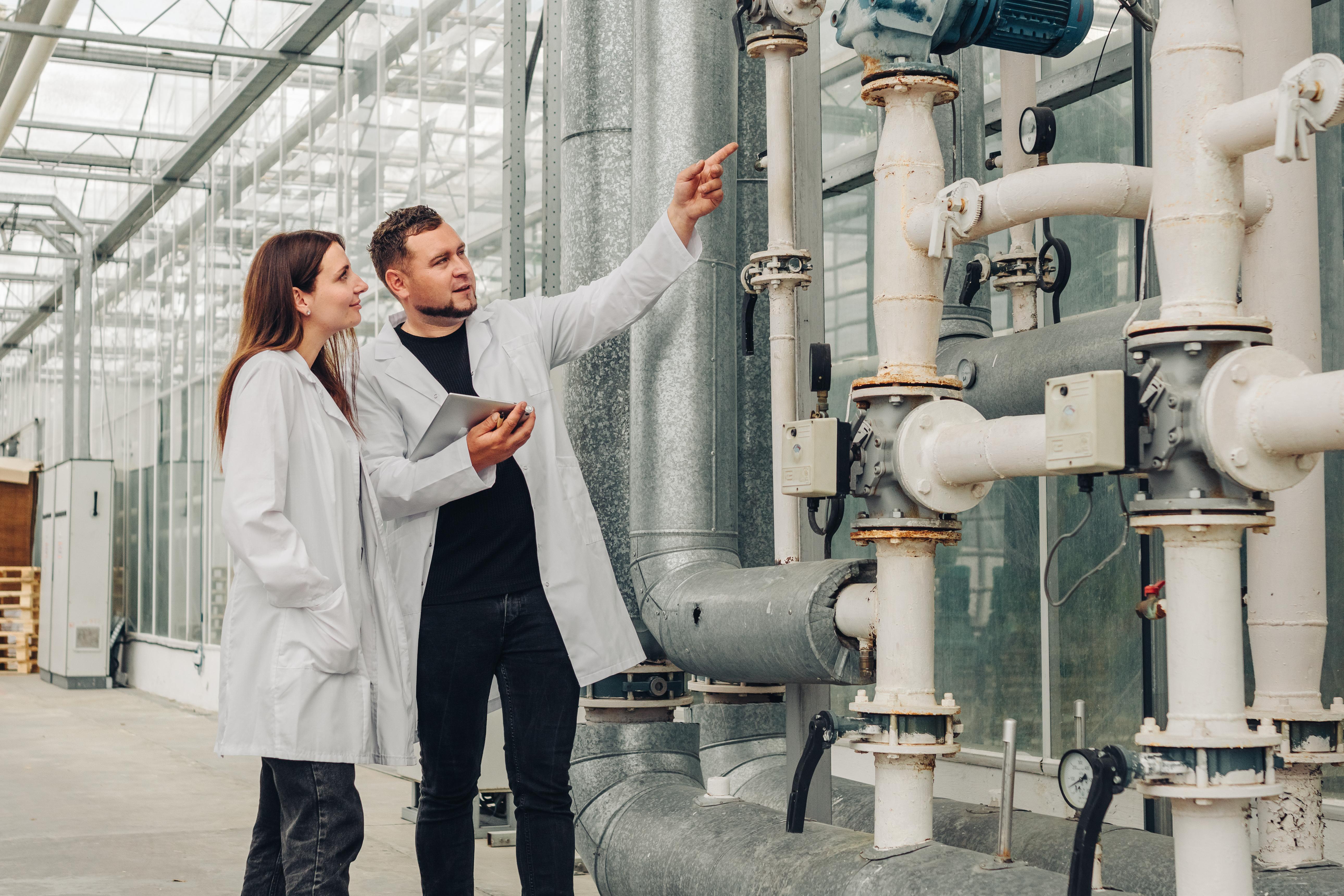 Two scientists in white lab coats inspecting a system of industrial pipes inside a greenhouse.