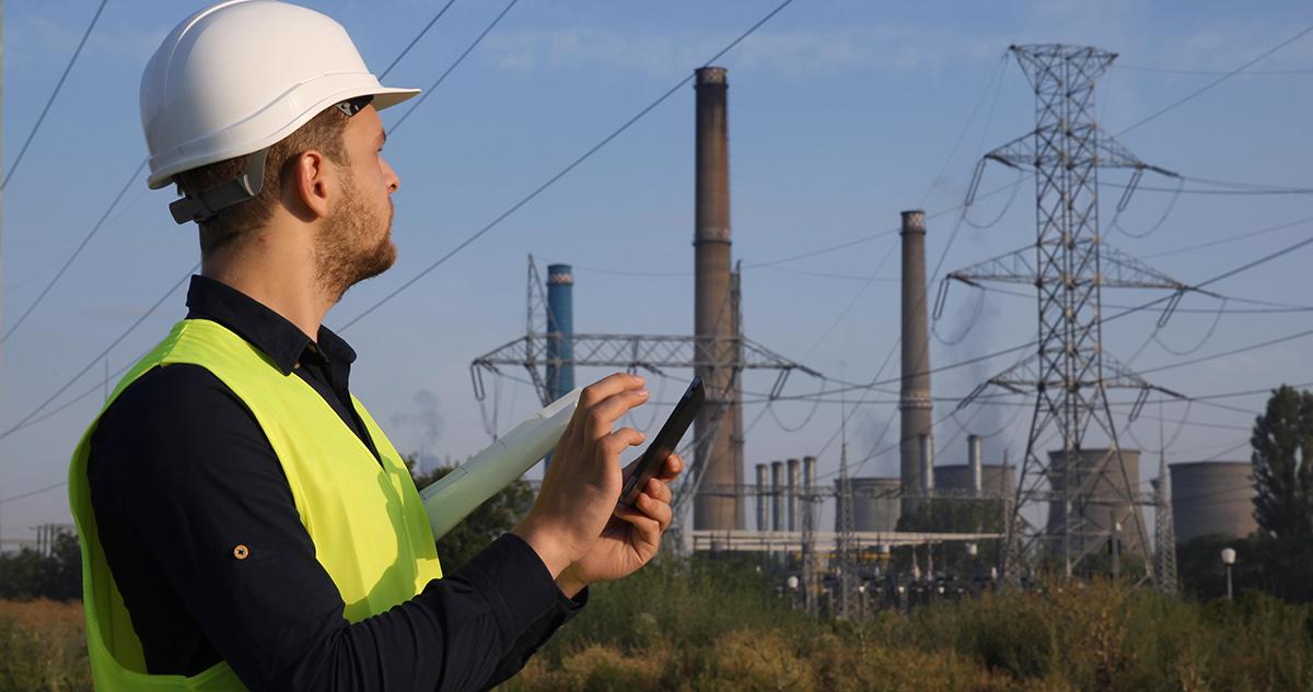 An engineer in a hard hat and safety vest using a tablet at an electrical power plant.